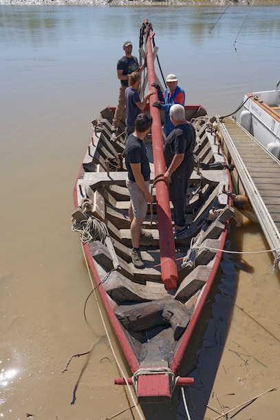 Le mât est à bord. Il faut maintenant le positionner sur son emplanture située juste derrière le banc de nage. A la poupe, une croix de saint André a été installée temporairement pour recevoir le mât en position couchée. © www.tessier.photos