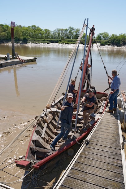 À l'avant de l'emplanture du mât, une chèvre a été positionnée et vissée sur une membrure. L'écoute de vergue est nouée à l'extrémité haute de la chèvre. Le mouflage relie le haut de la chèvre au quai. Il n'y a plus qu'à souquer et le mât se redresse enfin ! © www.tessier.photos